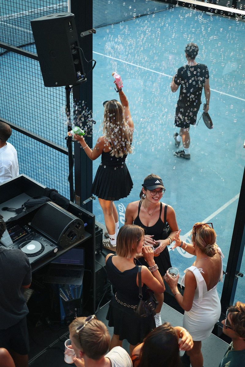 DJ-Pult direkt am Court bei Padel Parranda München: Gäste mit Seifenblasen davor, dahinter wird weiter Padel gespielt