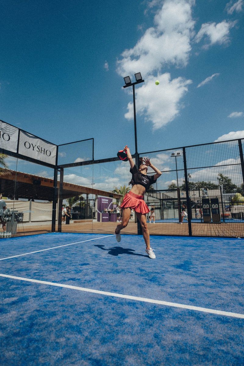 Spielerin springt auf dem blauen Padel-Court zum Überkopfschlag, darüber blauer Himmel, am Glas OYSHO-Banner